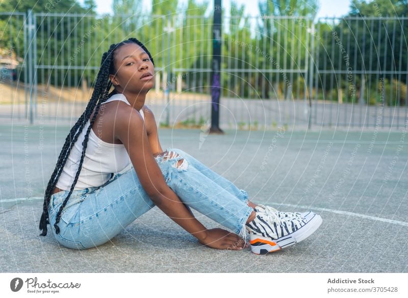 Junge schwarze Frau sitzt auf Sportplatz urban Stil Streetstyle trendy Hipster jung Sportpark Geflecht ethnisch tausendjährig Jeanshose Basketball Outfit Mode