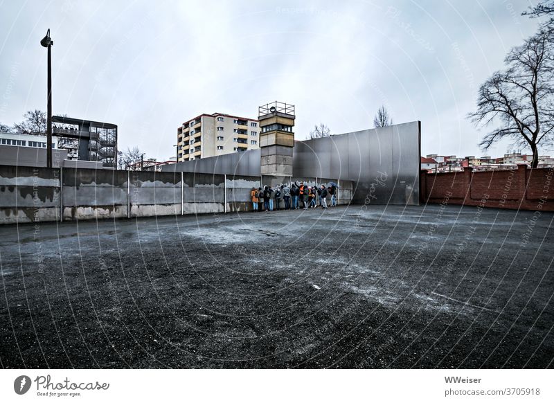 Eine Gruppe junger Touristen auf Tuchfühlung mit der Berliner Mauer Mauerstreifen Gedenkstätte Denkmal Memorial nah Schotter Fläche leerer Vordergrund bewölkt