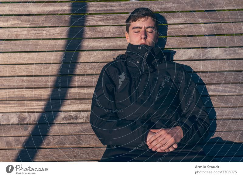 Junger Mann sitzt mit geschlossenen Augen auf einer Bank in der Sonne jung zugeklappt Sitzen Sonnenbad Sonnenlicht im Freien schlafen sich[Akk] entspannen
