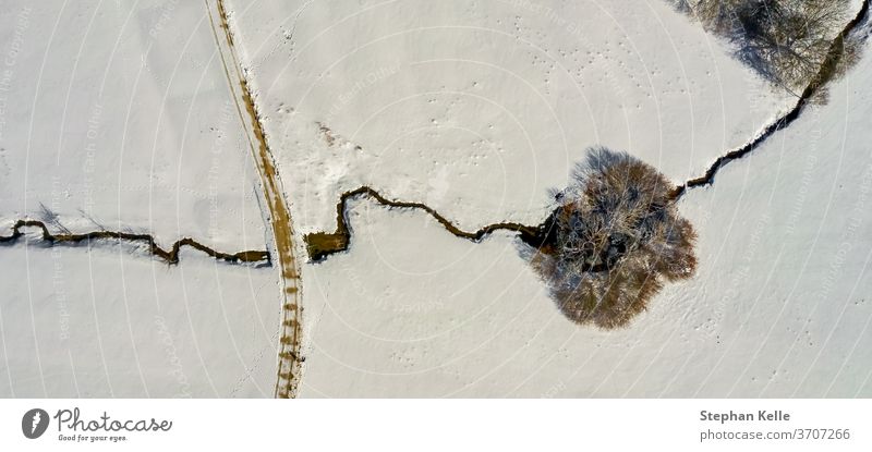 Winterlandschaft: Ein einsamer Baum, der von einem kleinen Bach durchquert wird, Landschaft voller Schnee. Fluss Single Straße allein Textfreiraum Dröhnen