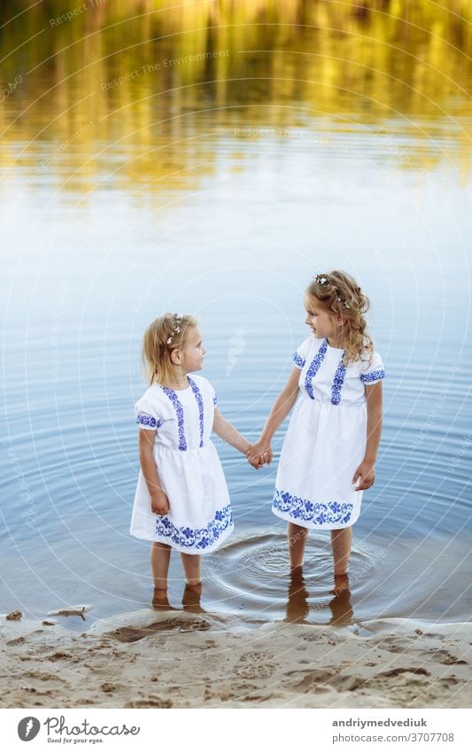 Zwei bezaubernde kleine Schwestern, die an einem warmen und sonnigen Sommertag lachen und sich umarmen. zwei Schwestern in weißen Kleidern in der Nähe des Wassers.