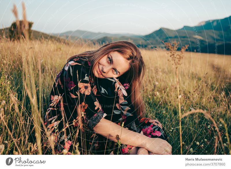 Glückliche Frau sitzt im Grasfeld mit Bergen im Hintergrund im Sonnenuntergang Abenteuer schön Schönheit braun Kaukasier niedlich Kleid Feld Blume Freiheit