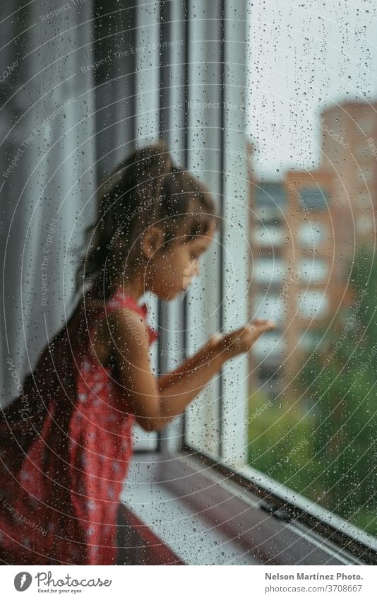 Kleines Mädchen, das an einem regnerischen Tag durch das Fenster in ihrem Schlafzimmer schaut. Sie steht auf einem weißen Stuhl, rückwärts gewandt. Wir können Tropfen im Kristall sehen.