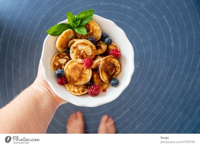 Hand mit frischen kleinen Pfannkuchen in einer weißen Schüssel. Frische Beeren, Draufsicht. Pancakes halten schüssel Barfuß blau fußboden Frühstück gebraten