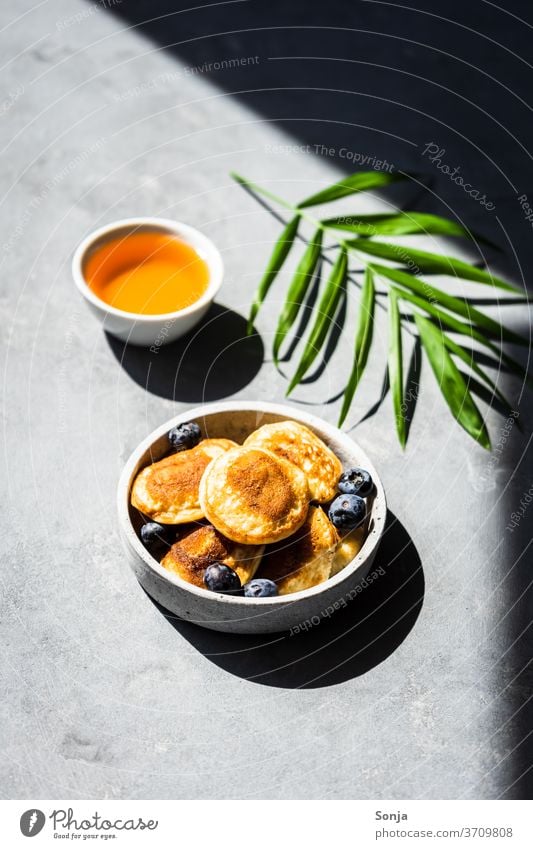 Mini Pfannkuchen mit frischen Heidelbeeren und Honig auf einem grauen Hintergrund. Schlagschatten und Licht. Pancakes klein Frühstück gebraten heiß zubereitet