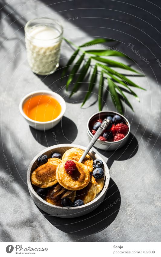 Mini Pancakes mit frischen Beeren und Honig auf einem grauen Tisch. Schlagschatten und Licht, Kontrastreich. Frühstück Dessert süß Pfannkuchen gebraten