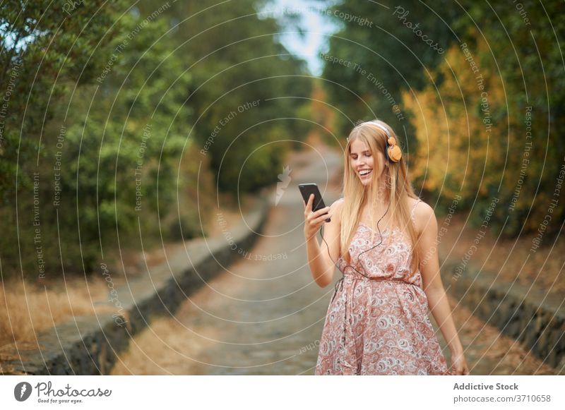Lächelnde Frau mit Smartphone und Kopfhörer zu Fuß im Park Glück zuhören benutzend Musik Sommer genießen jung Browsen Gasse Weg Gerät Apparatur Lifestyle Stil