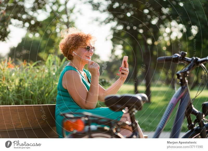 Reife Frau nutzt Videoanruf auf ihrem Smartphone im Park 60s Lebensalter gealtert App Bank Fahrrad Anruf Kaukasier heiter Mitteilung Konnektivität Kopfhörer