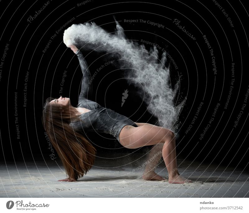Eine junge, schöne Frau mit langen Haaren trägt einen schwarzen Sportanzug, setzt sich auf den Boden und wirft weißes Mehl über sie zierlich Turner werfen