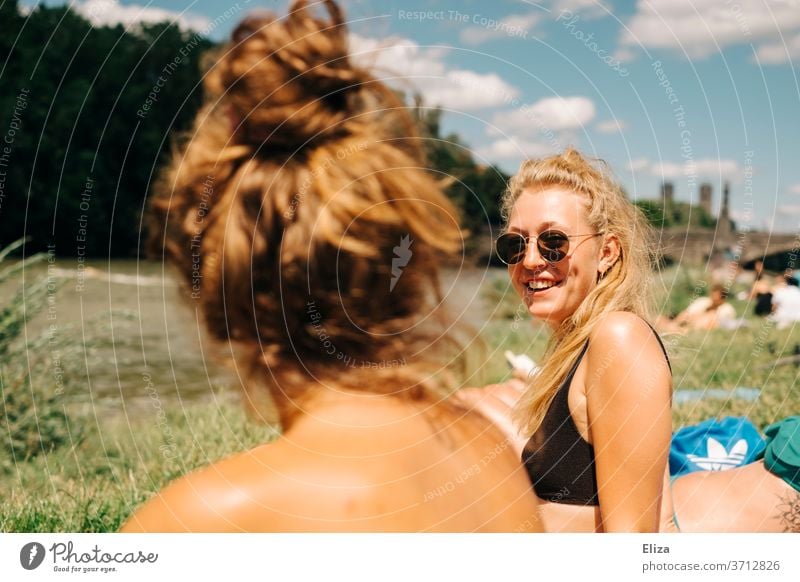 Zwei junge Frauen beim Sonnen und Baden an der Isar in München. Eine davon ist blond, trägt eine Sonnenbrille und lacht. Sommer. Freundinnen sonnen gute Laune