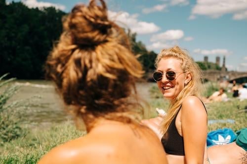 Zwei junge Frauen beim Sonnen und Baden an der Isar in München. Eine davon ist blond, trägt eine Sonnenbrille und lacht. Sommer. Freundinnen sonnen gute Laune