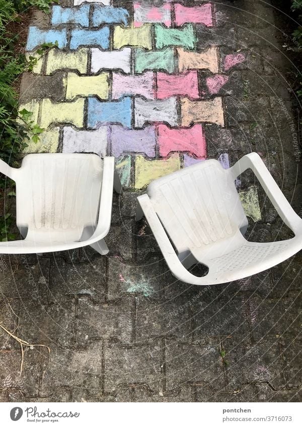 Zwei Gartenstühle aus Plastik für Kinder stehen vor einer bunten Malerei aus straßenkreide. Kinderspiel plastik kinderspiel straßenmalerei kreativität