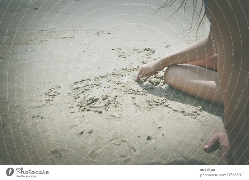 kind sitzt an einem strand und malt mit dem finger ein bild in den sand Kind Kindheit Mädchen Strand Sand Sommer Sommerferien am Meer Urlaub malen spielen Küste