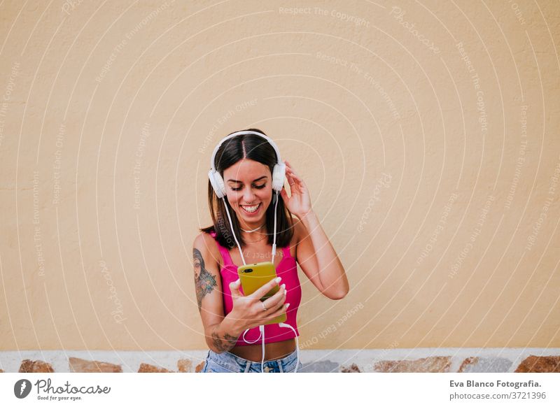 Fröhliche junge Frau im Freien, die mit Handy und Headset Musik hört. Technologie und Lebensstil Musik hören Glück Tattoo Technik & Technologie Großstadt urban