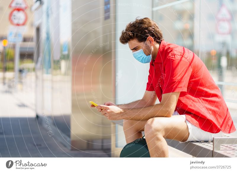 Junger Mann trägt eine chirurgische Maske, während er an einem Außenbahnhof auf einen Zug wartet Schüler Mundschutz Tourismus Ökomobilität Reisender jung