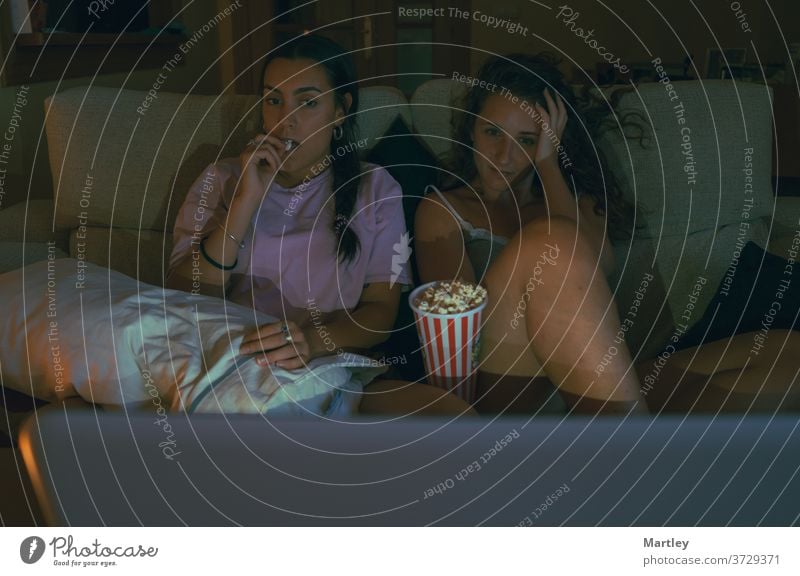 Zwei Teenager Mädchen essen Popcorn und schauen online einen Film an, während sie zusammen auf dem Sofa in einem gemütlichen Wohnzimmer sitzen. Popkorn