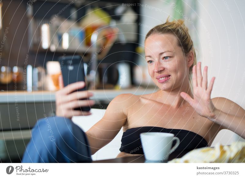 Junge, lächelnde, fröhliche, zufriedene Frau, die zu Hause in der Küche sitzt und soziale Medien auf dem Mobiltelefon nutzt, um mit ihren Lieben zu chatten und in Verbindung zu bleiben. Zu Hause bleiben, sozial distanzierender Lebensstil.