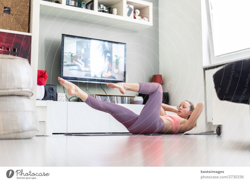 Attraktive sportliche Frau, die zu Hause trainiert und in ihrer kleinen Studiowohnung vor dem Fernseher Pilates-Übungen macht. Soziale Distanzierung. Bleiben Sie während einer Coronavirus-Pandemie gesund und bleiben Sie zu Hause
