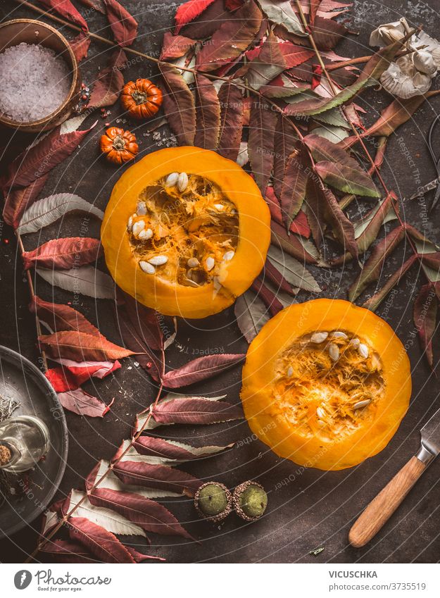 Saisonale Lebensmittel. Halber Hokkaido-Kürbis mit Samen, Messer und Herbstlaub auf dunklem Küchentisch. Herbstliche Stimmung. Ansicht von oben