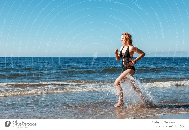 ein junges blondes Mädchen in einem schwarzen Badeanzug läuft am Sand des Meeresufers entlang Schönheit Kaukasier Körper schön Model Frau Wasser Person Sommer