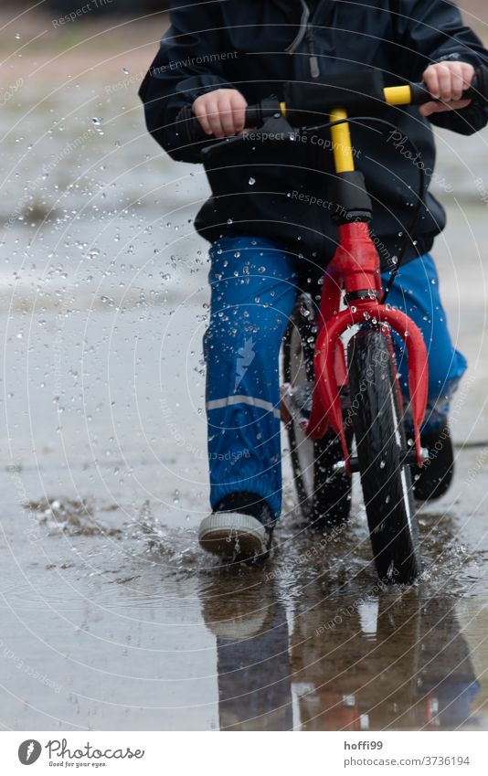 Kind auf Laufrad fährt mit Schwung durch die Pfütze Kinderspiel Kleinkind Pfützenbild spritzen Wassertropfen nass Spielen Kindheit Mensch Regen 1