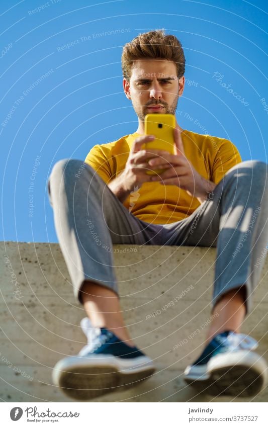 Mann benutzt sein Smartphone und sitzt draußen auf einer Kante männlich Lifestyle im Freien jung klug Telefon Typ Frisur Zelle Hintergrund modern lässig