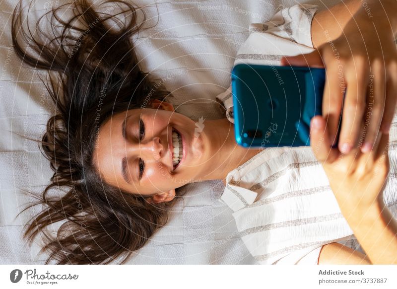Fröhliche Frau mit Smartphone auf dem Bett sich[Akk] entspannen heimwärts unterhalten Lächeln Talkrunde benutzend soziale Netzwerke Lügen weich Internet Browsen