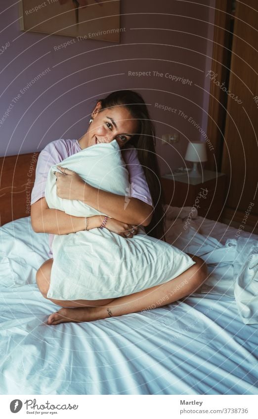 Lächelnde junge Frau in Schlafkleidung, die sich im Sitzen mit dem Kissen bedeckt und mit einem Arm am Kopf auf einem bequemen Bett liegt und sich morgens zu Hause entspannt.