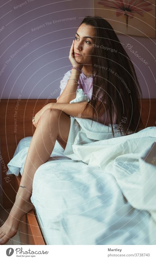 Gefällige Frau in Nachtwäsche, die ihr Gesicht berührt und zum Fenster schaut, während sie morgens auf einem weichen Bett sitzt. Menschen Erwachsener Porträt