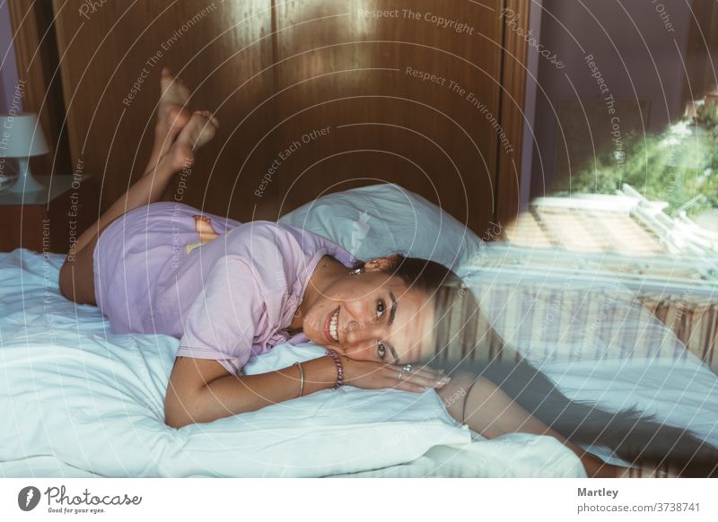 Porträt eines hübschen jungen Mädchens, das morgens in die Kamera schaut und lacht und auf einer weißen Bettdecke liegt, durch Glas fotografiert. sexy Lippe