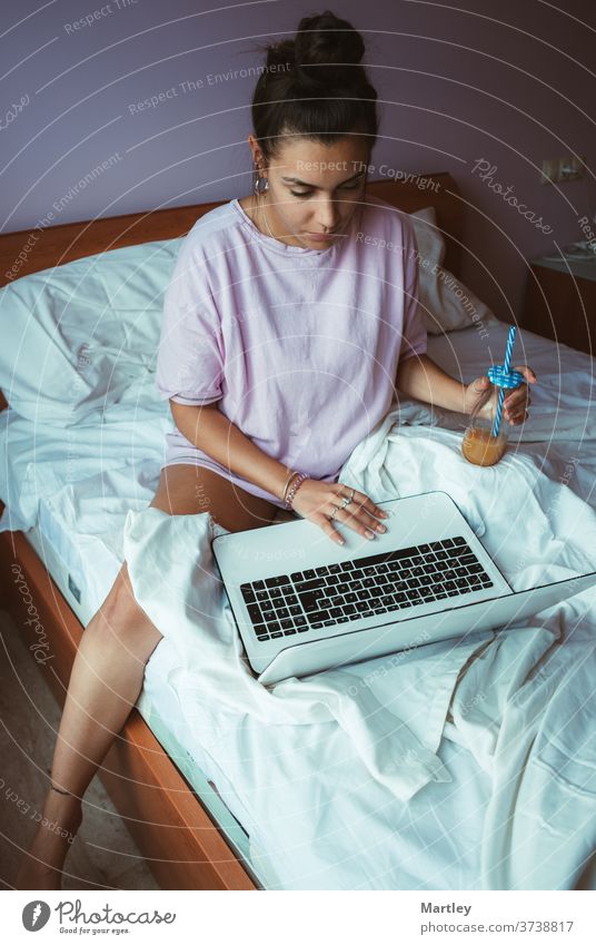 Barfüßige junge Frau im Pyjama, die Telearbeit leistet und erfrischenden Saft genießt, während sie morgens auf einem weichen Bett sitzt und am Laptop stöbert.