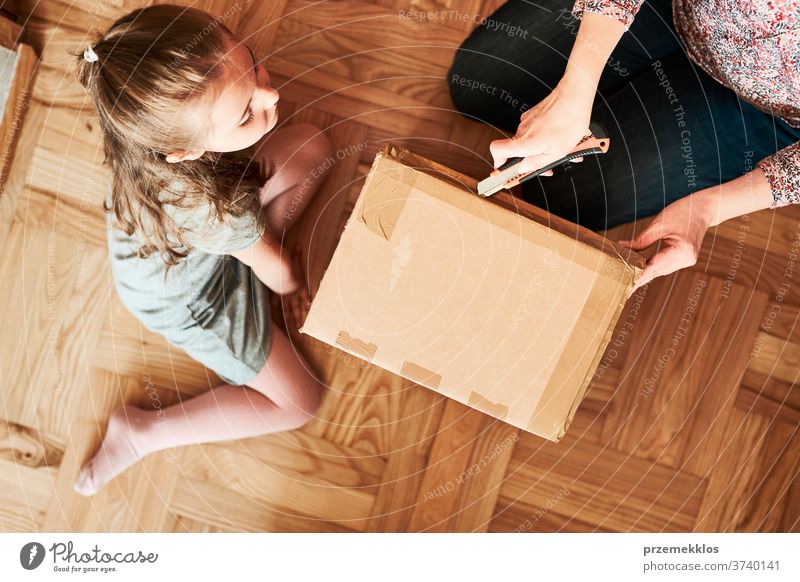 Frau beim Auspacken eines Pappkartonpakets im Zimmer zu Hause. Kleines Mädchen wartet auf das Öffnen eines Geschenks im Paket Ansicht Stock Klebeband Tochter