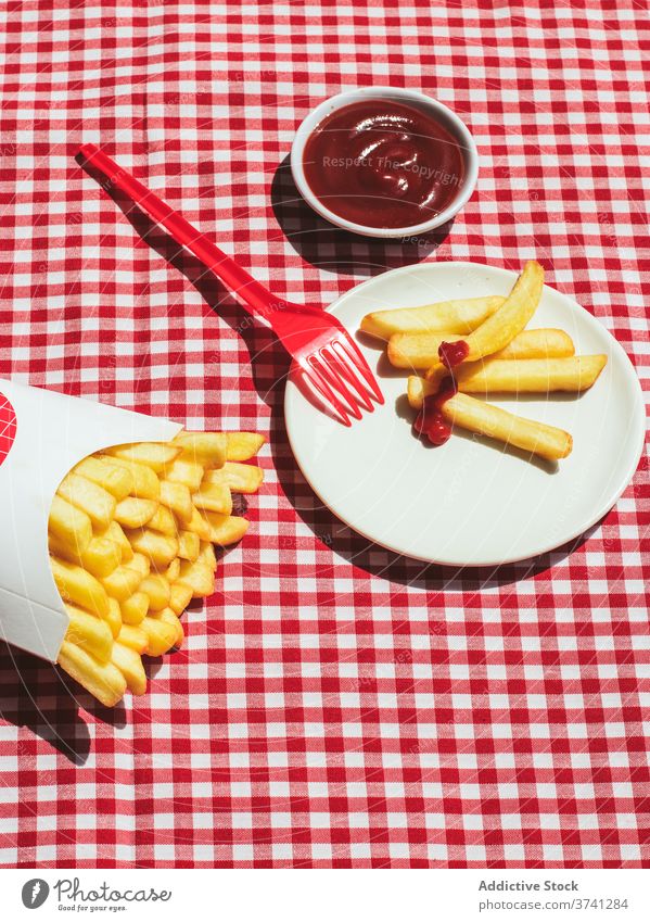Pommes-Paket neben Teller mit in Ketchup getränkten Kartoffeln Amerikanische Kultur Gewürz Kartoffelchips ungesunde Ernährung roter Hintergrund weiß Fritten