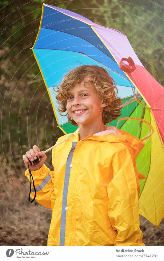 Junge im Regenmantel hält einen Regenschirm und lächelt in die Kamera Schuljunge gerade Pose Dusche Einstellung Regenbogen fallend Halt Gießen nass Wetter