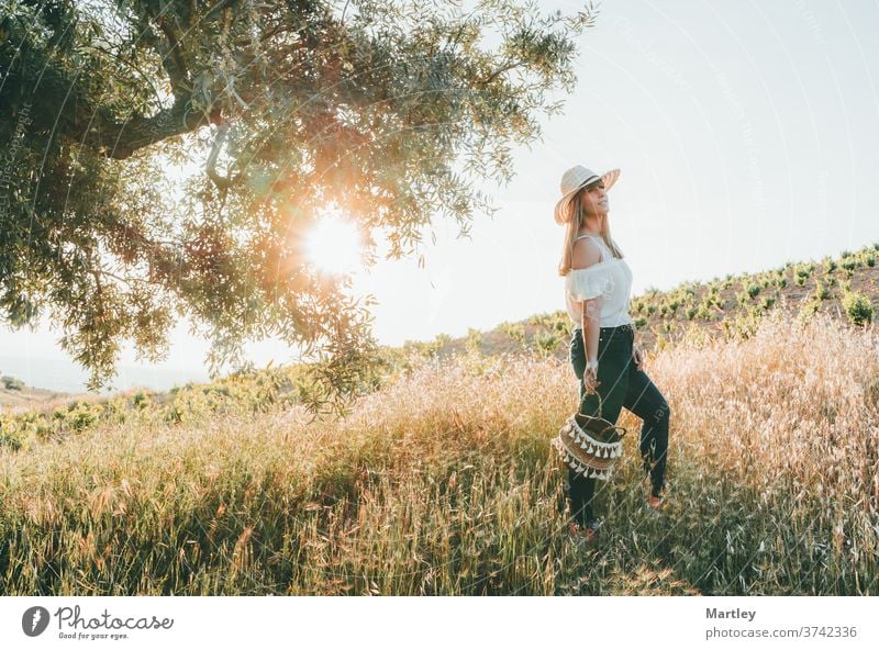 Junge Frau mit Strohhut und einem Korb, die im Sommer bei Sonnenuntergang durch das Feld geht. Konzept von Freiheit, Ruhe, Natur, Ökologie, grüner Welt und Glück.