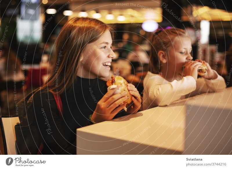 Hübsches Schulmädchen isst Hamburger und sitzt drinnen in einem Cafe. Glückliches Kind, das Junkfood in einem Restaurant isst. Seitenansicht Lebensmittel