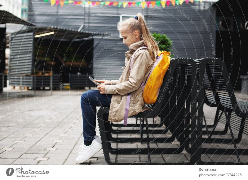 Nach der Schule sitzt ein süßes Mädchen auf einer Bank und wartet auf den Bus, lernt online auf einem Tablett und kommuniziert per Video. Tablette Rucksack