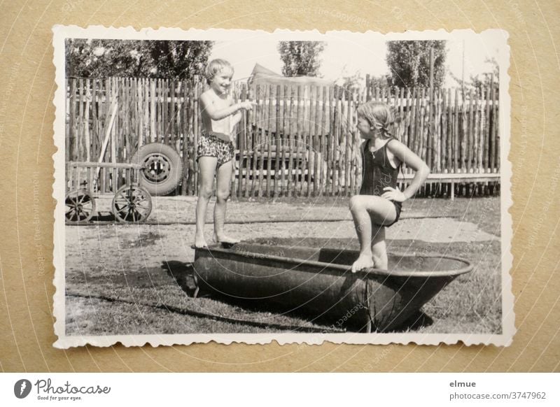 Erinnerungen an die 1960er Jahre - ein schwarz-weißer Fotoabzug mit Büttenrand liegt auf beigefarbenem Papier und zeigt zwei Mädchen beim Baden in einer alten Zinkbadewanne in ländlicher Umgebung mit Zaun, Handwagen und Autoreifen