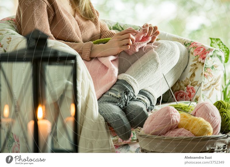 Junge Frau sitzt zu Hause auf einem Stuhl am Fenster, trägt einen gestrickten warmen Pullover und strickt mit Nadeln. Gemütliches, mit Laternen und Kerzen dekoriertes Zimmer. Szenische Winteransicht von Kiefern im Schnee im Fenster