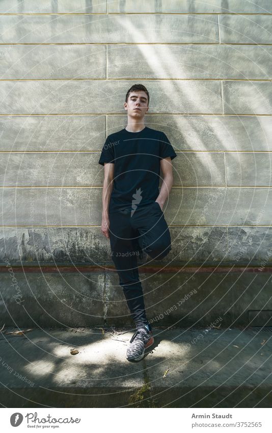 Porträt eines kühlen, stehenden jungen Mannes, der an eine Wand gelehnt ist Stehen Lehnen Grunge selbstbewusst ernst Sportbekleidung Schmutz im Freien Teenager