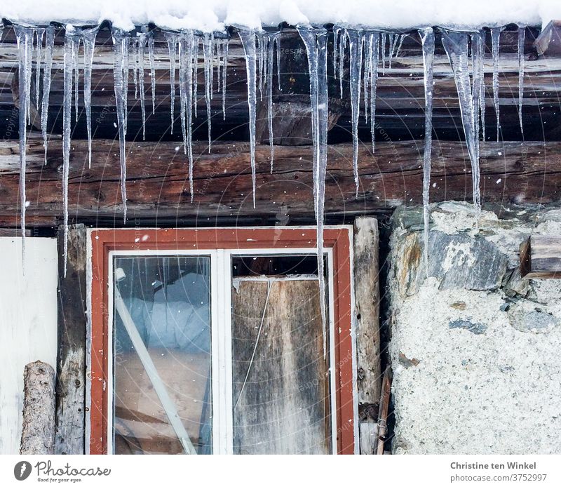 Lange Eiszapfen hängen vom Dach einer alten Hütte. Detailansicht mit rotem Fensterrahmen, Steinen und Holzbalken Frost Winter Winterstimmung Steinwand Eiszeit