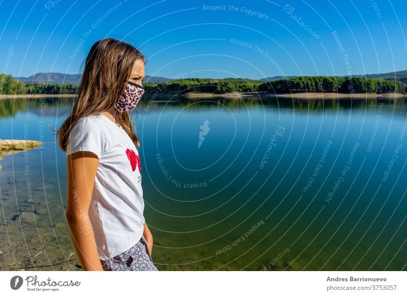 Mädchen mit weissem T-Shirt und einer Maske zum Schutz vor dem Coronavirus, das in einem See posiert, in dem sich der Wald im Wasser spiegelt niedlich jung