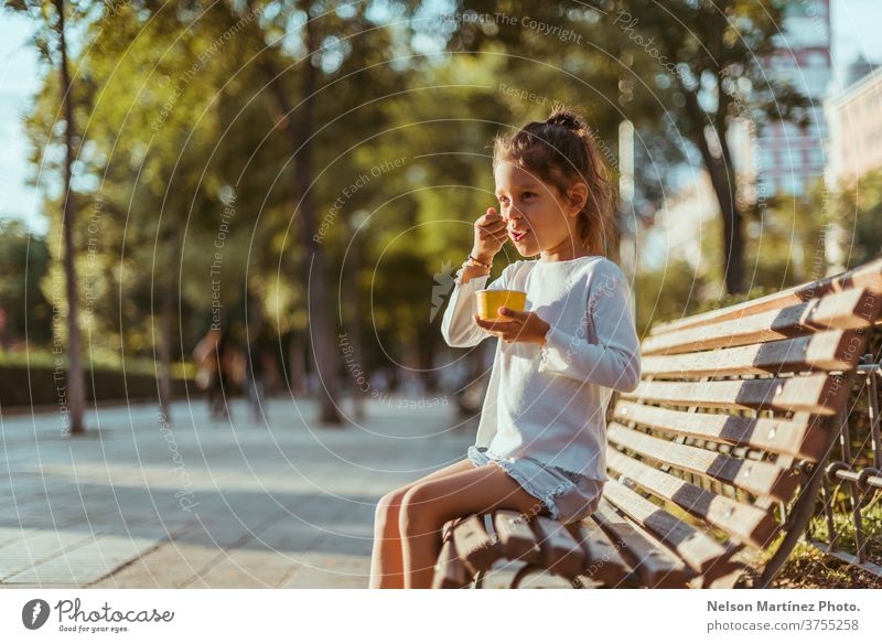 Lateinamerikanisches Kind, das auf einer Parkbank mit schönen Bäumen im Hintergrund sitzt. Sie isst gerade ein Eis. außerhalb wirklich Glück Sonnenuntergang