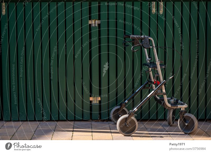 Einsamer Rollator vor grüner Holzwand Gehhilfe Senior Außenaufnahme alt Mobilität Handicap Hilfsmittel mobil Gesundheit Wand Einsamkeit Pflege textfreiraum