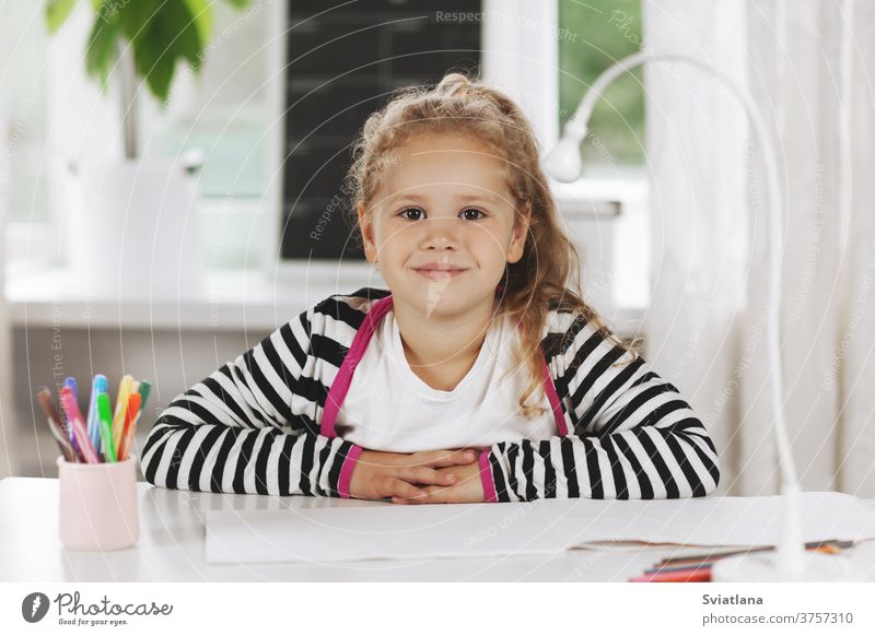 Porträt eines charmanten kleinen Mädchens, das an einem Tisch sitzt. Das Mädchen sitzt am Tisch, auf dem Tisch liegt ein Skizzenbuch, Buntstifte. Hausaufgaben, Hausunterricht, soziale Distanz