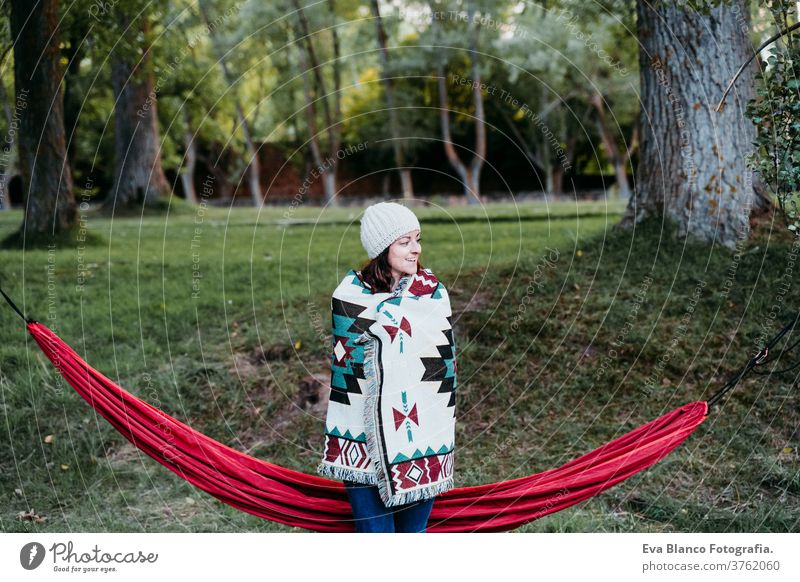 junge Frau, die mit der Decke bedeckt, die nahe bei der Hängematte steht. Herbstsaison. Camping-Konzept kalt im Freien Natur Sonnenuntergang orange Park