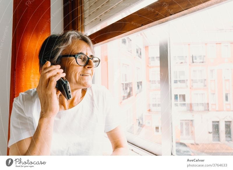 Nahaufnahme einer alten Frau, die mit dem Handy telefoniert und lächelnd durch das Fenster schaut Großmutter im Innenbereich Ruhestand Person Mitteilung