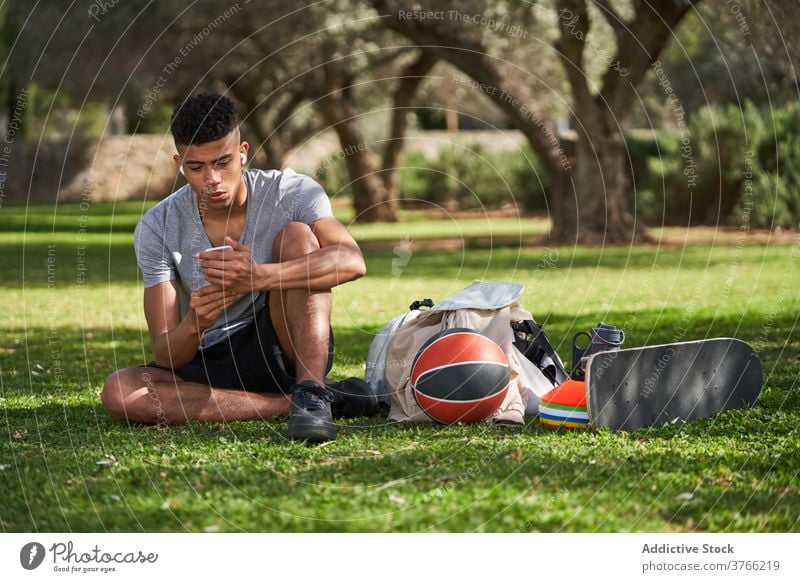 Schwarzer sportlicher Mann sitzt auf Rasen und benutzt Smartphone Sportler Basketball sich[Akk] entspannen Training Talkrunde Athlet Sommer männlich ethnisch