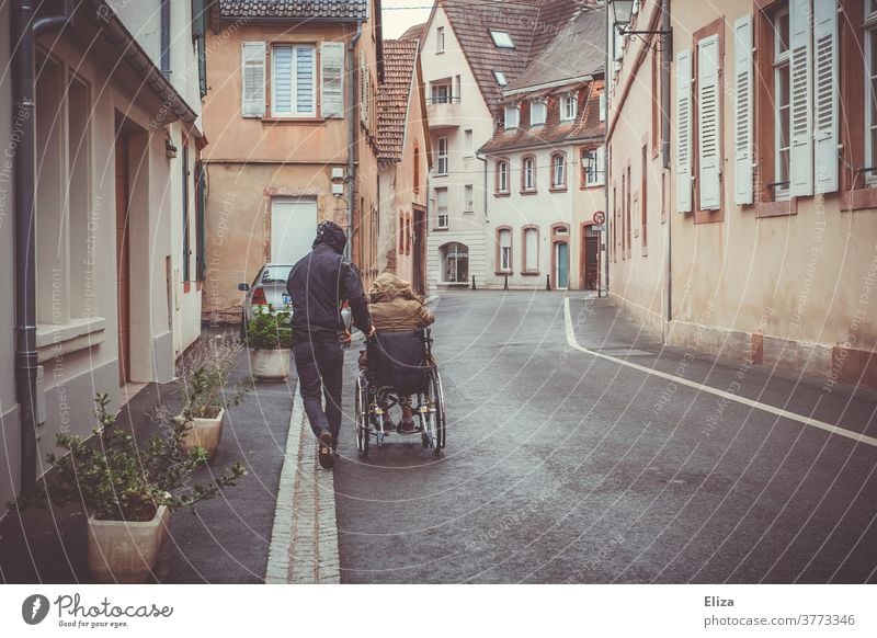 Ein Mann schiebt eine Person mit Behinderung im Rollstuhl die Straße entlang Mobilität Handicap Gesundheitswesen Pflege Regenwetter Krankheit Ausflug schieben
