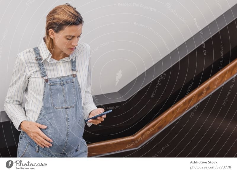Schwangere Frau beim Surfen auf dem Smartphone in einem modernen Gebäude schwanger benutzend Nachricht Bauch mütterlich Schwangerschaft Zeitgenosse Jeansstoff